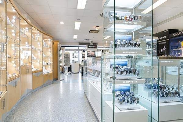 Das Innere eines Uhrenladens mit Glasvitrinen auf beiden Seiten, Reihen von Uhren im Schaufenster, Holzschränken und einem leeren Gang.