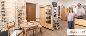 Ein Optikerladen mit einer stehenden Frau. Verschiedene Brillen sind ausgestellt, zusammen mit Stühlen und einem Tisch. Der Laden hat Holzschränke und eine weiße Wand.