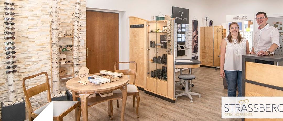 Ein Optikerladen mit einer stehenden Frau. Verschiedene Brillen sind ausgestellt, zusammen mit Stühlen und einem Tisch. Der Laden hat Holzschränke und eine weiße Wand.