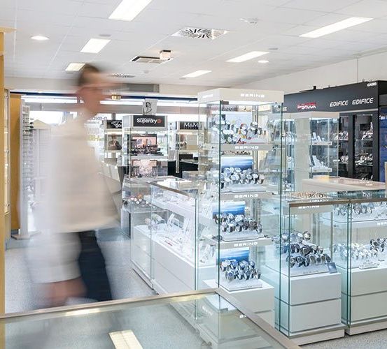 Ein Juwelierladen mit vielen Glasvitrinen, die mit Uhren und Schmuck gefüllt sind. Der Laden hat ein modernes Design mit weißen Regalen und Glasdisplays. Zwei Personen gehen im Laden.