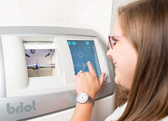 Eine Frau mit Brille und Uhr interagiert mit einem grauen medizinischen Gerät mit der Aufschrift 'briot'. Sie drückt auf den Bildschirm und lächelt.