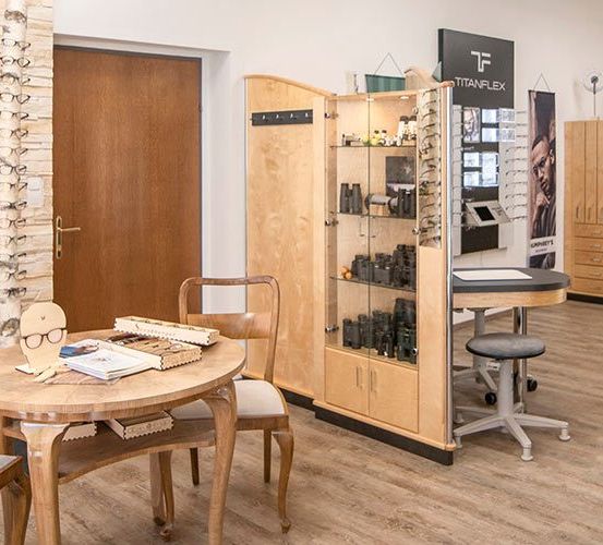 Ein Optikgeschäft mit Holzmöbeln, einem Tisch mit einem Buch und einer Ausstellung von Brillen. Eine Frau steht vor einer Wand mit Regalen und einem Schild.