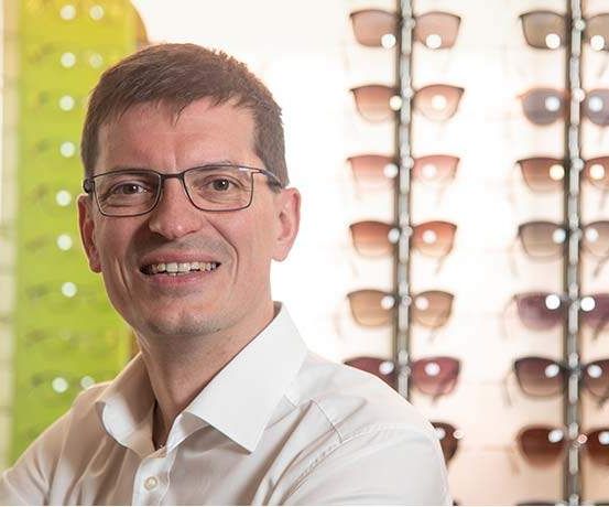 Ein Mann mit Brille lächelt und steht vor einer Wand mit einer Ausstellung von Sonnenbrillen.