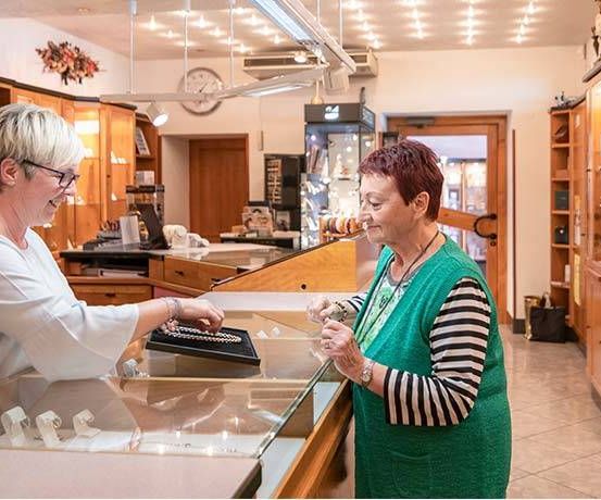 Zwei Frauen stehen bei einer Schmuckvitrine, lächeln und besprechen Schmuck. Im Hintergrund befinden sich eine Uhr und ein Regal mit verschiedenen Gegenständen.