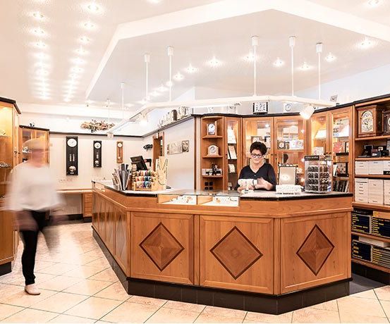 Eine Frau steht hinter einer hölzernen Schmucktheke, umgeben von Regalen mit Uhren und Schmuck. Eine andere Person geht vorbei. Der Raum hat Holzschränke, Fliesen und Deckenleuchten.