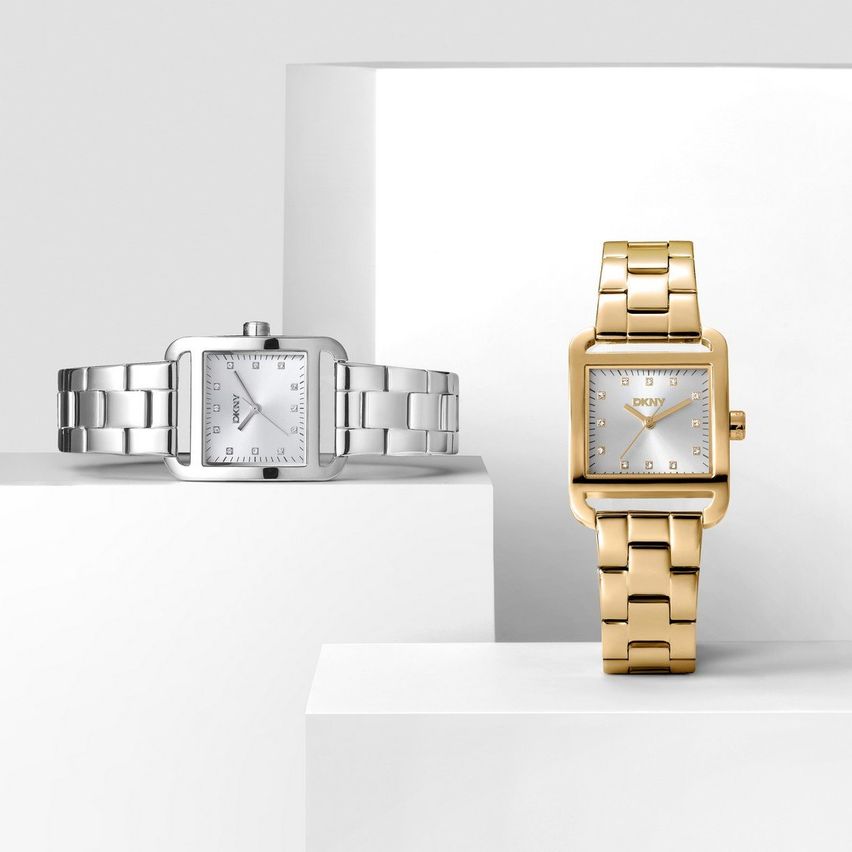 Zwei DKNY Uhren, eine in Gold und eine in Silber, sind auf weißen Oberflächen ausgestellt. Beide haben quadratische Zifferblätter mit analogen Anzeigen.