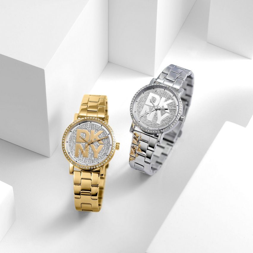 Zwei DKNY-Uhren, eine goldfarbene und eine silberne, sind auf einer weißen Oberfläche ausgestellt. Die goldene Uhr hat ein funkelnde Gesicht mit 'DKNY' in Gold. Die silberne Uhr hat ein diamantbesetztes Gesicht mit 'DKNY' in Weiß.