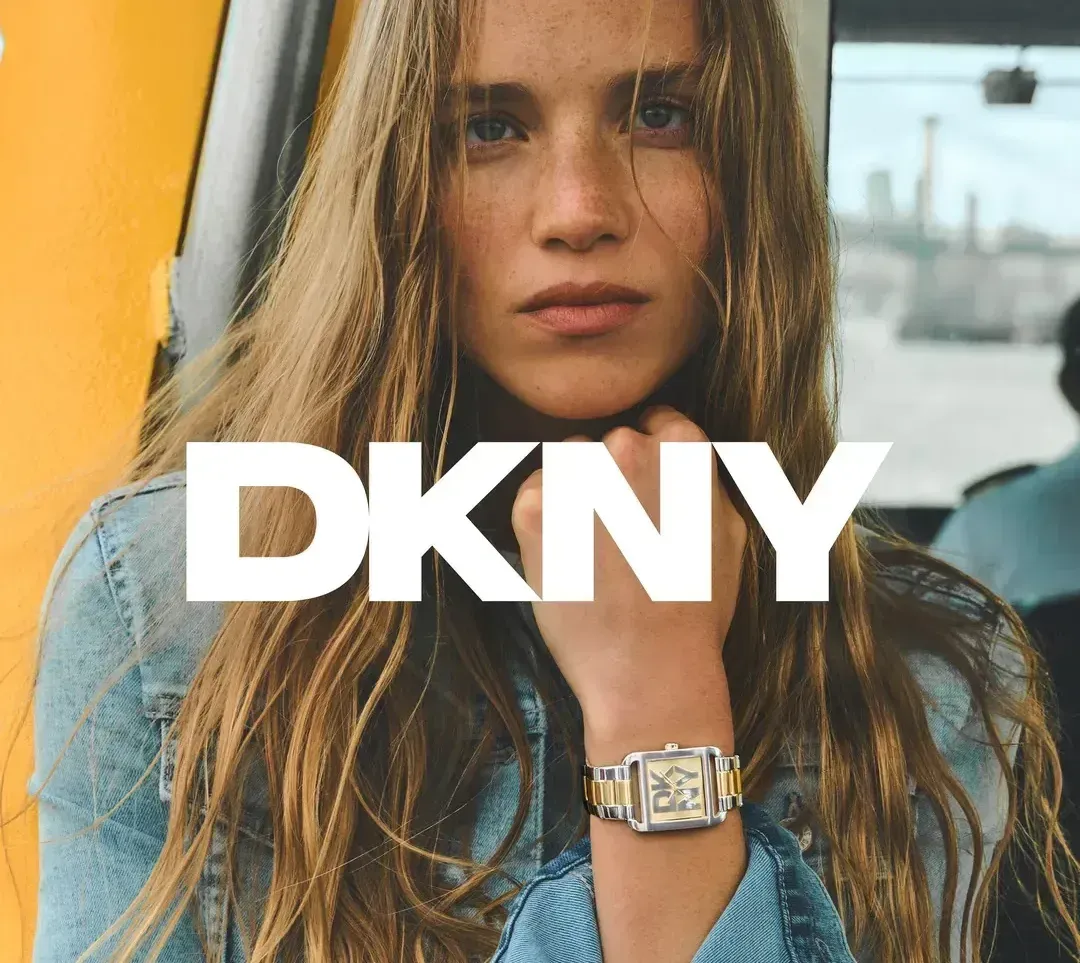 Eine Frau mit langen Haaren trägt eine Jeansjacke und eine DKNY-Uhr. Der Hintergrund ist unscharf mit einer gelben Wand und Gebäuden.