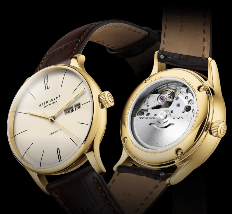 Zwei goldfarbene Uhren mit Lederarmbändern werden gezeigt. Die linke Uhr hat ein weißes Zifferblatt und den Text 'STERNGLAS ZEITMESSE'. Die rechte Uhr zeigt ihre inneren Mechaniken, gekennzeichnet mit 'MIYAOTA JAPAN'.