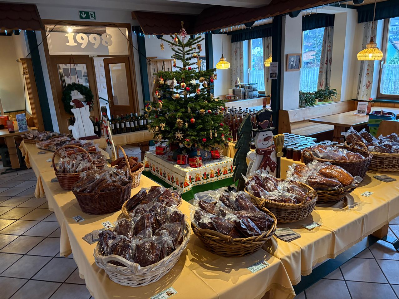 Ein Raum mit verschiedenen Körben gefüllt mit verpackten Lebensmitteln. Es gibt einen Weihnachtsbaum mit Dekorationen, einen Schneemann und einen Tisch mit Flaschen. Der Raum hat einen gefliesten Boden und Glasfenster.