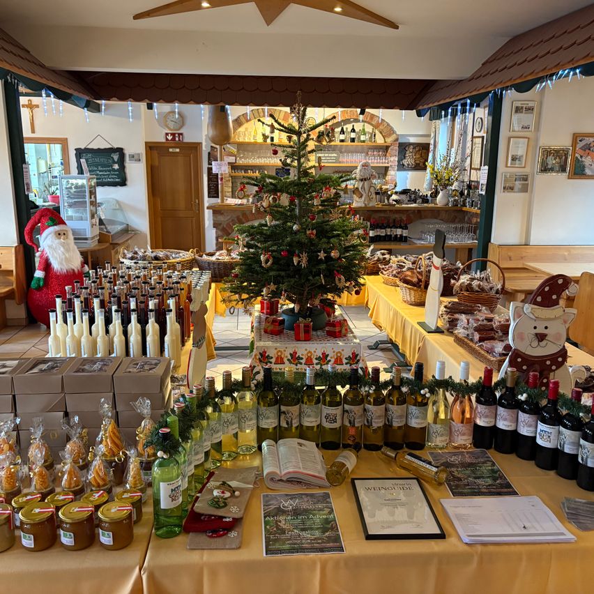 Ein Raum gefüllt mit verschiedenen Produkten und Geschenken, ein Weihnachtsbaum in der Mitte und eine Santa-Claus-Figur auf der linken Seite. Zu den Produkten gehören Flaschen Wein, Gläser und Bücher. Ein Schild und ein Kalender stehen auf dem Tisch.