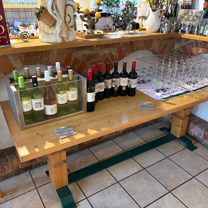 Eine Holzbank zeigt Weinflaschen und Gläser. Eine Weinbox hält weitere Flaschen. Eine Ziegelwand und ein Regal mit verschiedenen Gegenständen sind im Hintergrund.