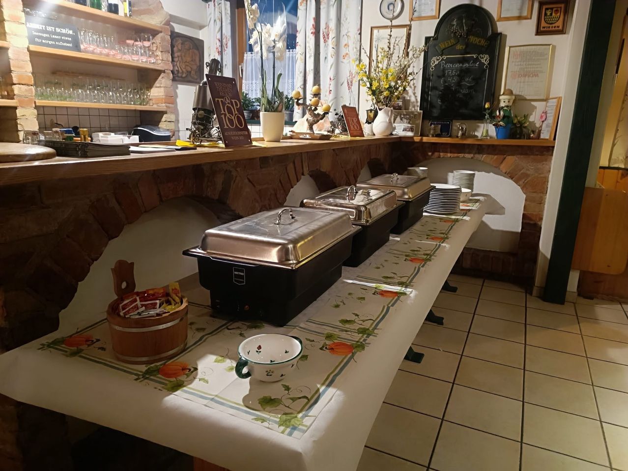A long table with a tablecloth is set up in a restaurant with three chafing dishes and a bowl. The shelves have glasses and other items. There are flowers and plants in pots.