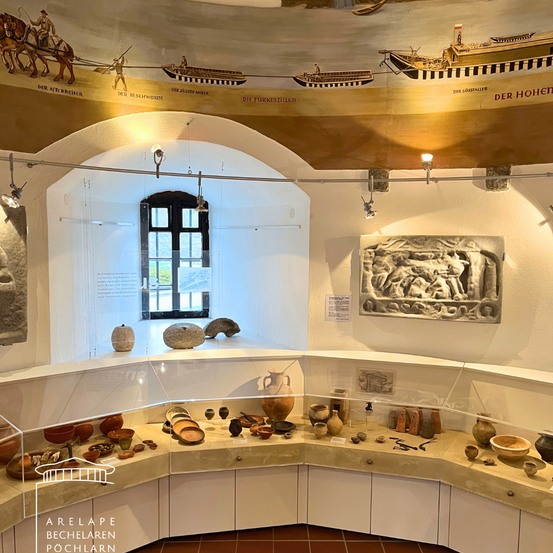 Ein Museum zeigt Keramik, Skulpturen und Artefakte, mit einem gewölbten Deckenbogen und einem Fenster. Boote sind an der Wand abgebildet und eine antike Reliefwand ist zu sehen.