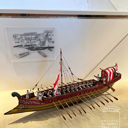 Ein Modell eines antiken Schiffes ist in einem Museum ausgestellt, mit einer Zeichnung einer Hafenszene an der Wand dahinter. Das Schiff hat Ruder und eine Segel, und es gibt Figuren von Menschen darauf.
