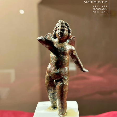 Eine bronzene Statue eines geflügelten Kindes steht auf einer weißen Basis. Sie ist in einem Museum ausgestellt, mit einer weißen Wand im Hintergrund.