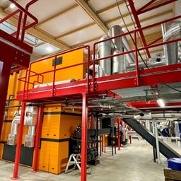 Eine industrielle Anlage im Innenbereich mit großen gelben und schwarzen Maschinen, die durch Rohre verbunden sind, unter einem Holzdach und einem roten Metallrahmen auf der linken Seite.