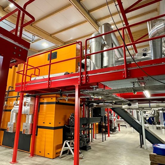 Eine industrielle Anlage im Innenbereich mit großen gelben und schwarzen Maschinen, die durch Rohre verbunden sind, unter einem Holzdach und einem roten Metallrahmen auf der linken Seite.