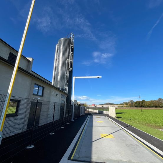 Ein Gebäude mit einem hohen Silo steht hinter einem Zaun, und eine Straße führt zu einem Tor mit einer Metallstruktur. Bäume und ein Feld sind in der Ferne.