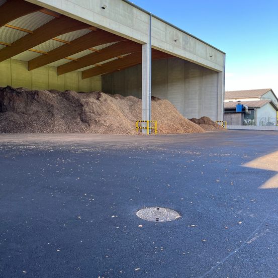 Ein großer Haufen aus trockenen Blättern und Holzspänen befindet sich vor einem großen Gebäude mit Metalldach und Betonwänden. Das Gebiet ist von gelben Barrieren umgeben.