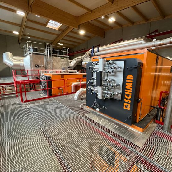 Eine Indoor-Anlage zeigt Industriemaschinen, darunter eine große orange-schwarze Einheit mit der Aufschrift SCHMID, umgeben von verschiedenen Rohren und Metallstrukturen.