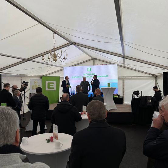 Eine Veranstaltung findet unter einem weißen Zelt mit einer großen Leinwand statt. Drei Männer stehen auf der Bühne mit Mikrofonen, einer hält einen Blumenstrauß. Menschen sitzen vor der Bühne um Tische herum.