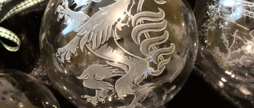 Mehrere Glasornamente sind auf einer schwarzen Oberfläche ausgestellt. Ein Ornament sticht besonders durch ein Drachendesign hervor. Der Drache ist detailreich dargestellt und auf einem klaren Glashintergrund gesetzt.