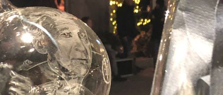 Eine Glasvase mit reflektierender Oberfläche und einem gravierten Gesicht steht auf einer schwarzen Oberfläche. Dahinter steht eine Person neben einem Weihnachtsbaum.