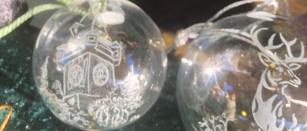 Zwei klare Glas-Weihnachtskugeln liegen auf einer grünen Oberfläche. Eine hat ein Glockenturmdesign und die andere ein Hirschdesign.
