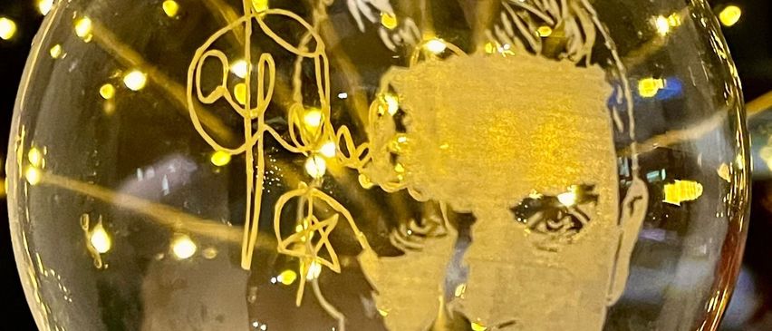 Eine Glaskugel mit einer goldenen Silhouette eines Männergesichts, mit einer Unterschrift, beleuchtet von gelben Lichtern im Hintergrund.