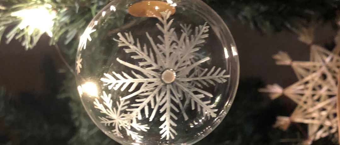 Ein Gläsernes Schneeflockenornament hängt an einem Weihnachtsbaum, erleuchtet von sanften Lichtern. Das Schneeflockendesign ist kunstvoll geätzt.