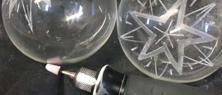 Drei Glaskugeln auf einer schwarzen Oberfläche. Zwei haben Sterndesigns und eine ist schlicht. Ein schwarzes Werkzeug mit einer rosa Spitze liegt neben ihnen.