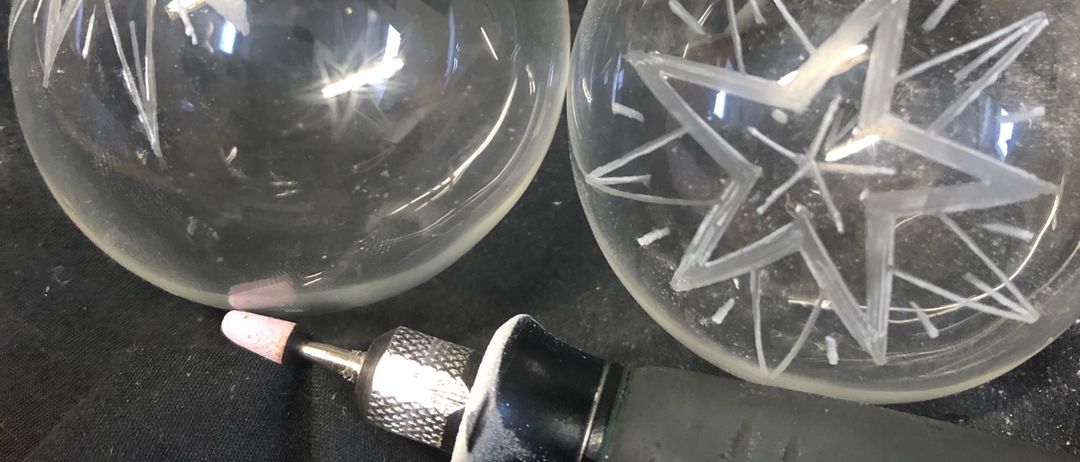 Drei Glaskugeln auf einer schwarzen Oberfläche. Zwei haben Sterndesigns und eine ist schlicht. Ein schwarzes Werkzeug mit einer rosa Spitze liegt neben ihnen.