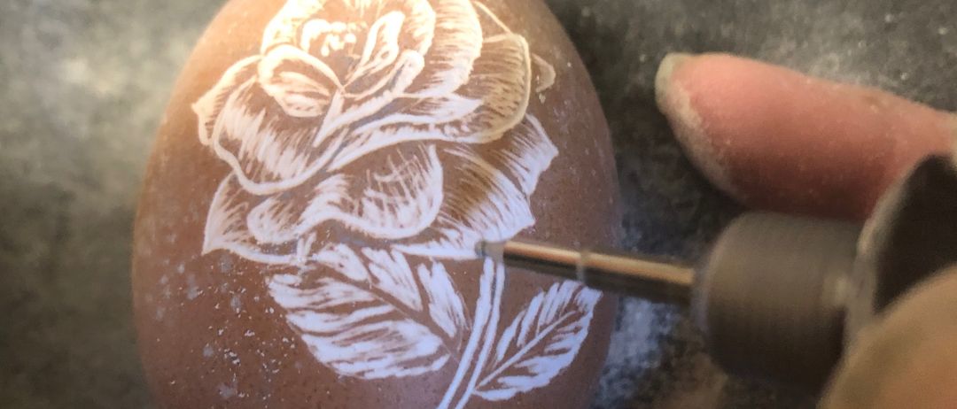 Eine Hand benutzt ein Werkzeug, um ein weißes Rosendesign in ein braunes Ei zu ätzen, mit einem Marmoruntergrund im Hintergrund.