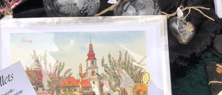 Auf einem Tisch sind verschiedene Glaskugeln, ein gerahmtes Bild und eine Karte mit einem Preisschild zu sehen. Das Bild zeigt eine Stadt mit einer Kirche und Bäumen.