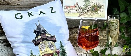 Ein dekoratives Kissen mit dem Wort Graz darauf, das einen Uhrturm zeigt, steht auf einem Tisch mit zwei Weingläsern, eines mit Zitronenscheibe und das andere mit einer Rose und Blättern.
