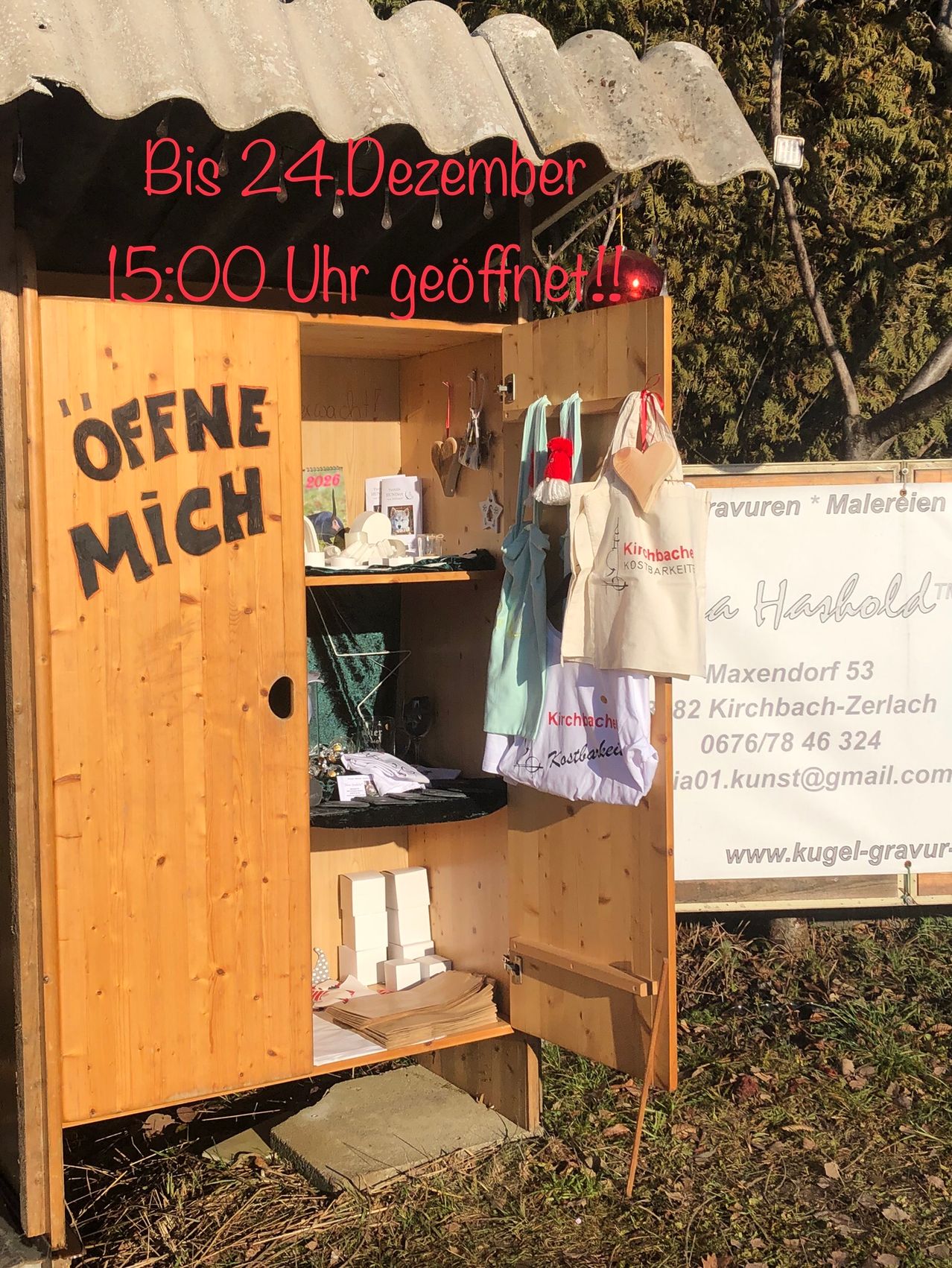 Ein hölzerner Schrank mit 'Offne Mich' beschriftet, geöffnet, um verschiedene Gegenstände zu zeigen. Rechts ein weißes Schild mit schwarzem Text. Hinter dem Schrank stehen Bäume und ein Gebäude.