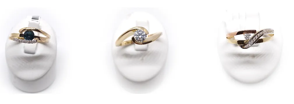 Drei goldene Ringe mit Diamanten sind auf weißen Oberflächen ausgestellt. Der mittlere Ring ist aus Gold mit einem großen Diamanten. Der linke Ring ist aus Gold mit einem kleineren Diamanten. Der rechte Ring ist eine Kombination aus Gold und Roségold mit zwei Diamanten.