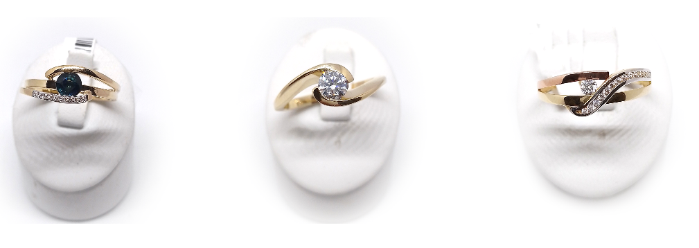 Drei goldene Ringe mit Diamanten sind auf weißen Oberflächen ausgestellt. Der mittlere Ring ist aus Gold mit einem großen Diamanten. Der linke Ring ist aus Gold mit einem kleineren Diamanten. Der rechte Ring ist eine Kombination aus Gold und Roségold mit zwei Diamanten.