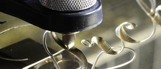 Eine Nahaufnahme einer Werkzeugmaschine mit komplizierten Metallarbeiten. Der metallische Körper der Maschine hat eine netzartige Textur und ein zylindrisches Metallstück. Die Metallarbeiten, einschließlich eines Rings und Spiralen, werden auf einer Glasoberfläche gefertigt.