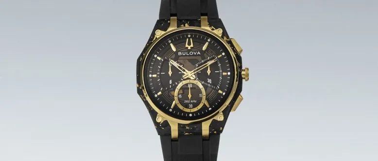 Eine schwarze und goldene Bulova Uhr mit rundem Zifferblatt, goldenen Akzenten und einem schwarzen Gummiband. Die Uhr liegt auf einem geschwungenen schwarzen Armband.