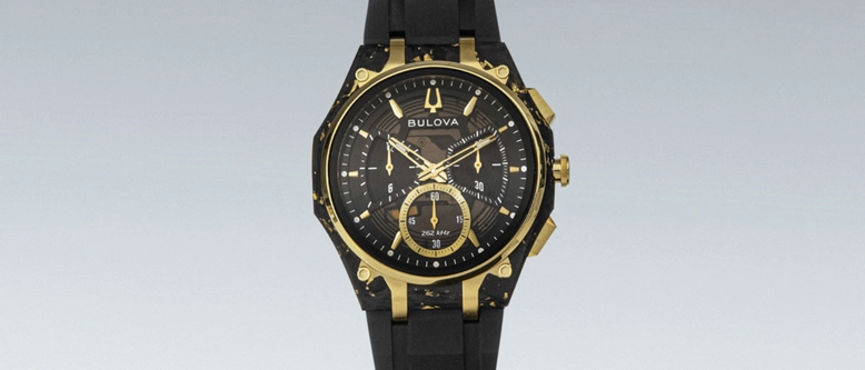 Eine schwarze und goldene Bulova Uhr mit rundem Zifferblatt, goldenen Akzenten und einem schwarzen Gummiband. Die Uhr liegt auf einem geschwungenen schwarzen Armband.