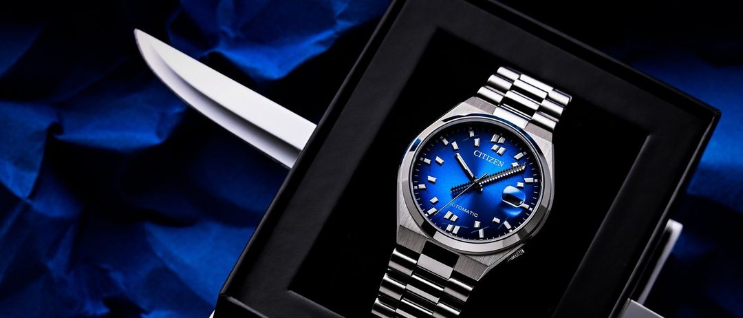 Eine Citizen Uhr mit blauem Zifferblatt liegt in einer schwarzen Schachtel auf einem blauen Tuch, mit einem Messer daneben.