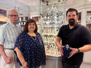 Drei Personen stehen vor einer Glasvitrine mit Uhren. Ein Mann auf der linken Seite trägt eine gestreifte Hemd und eine Brille. Eine Frau in der Mitte trägt ein blaues Kleid mit Punkten und eine Perlenkette. Ein Mann auf der rechten Seite hält eine Uhr in einer Schachtel.