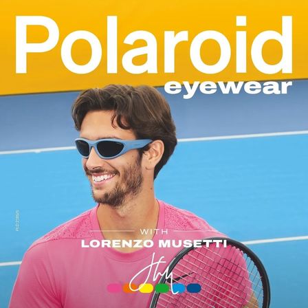 Ein Mann mit Polaroid-Brillen lächelt auf einem Tennisplatz. Er hält einen Tennisschläger und trägt ein rosa Shirt.