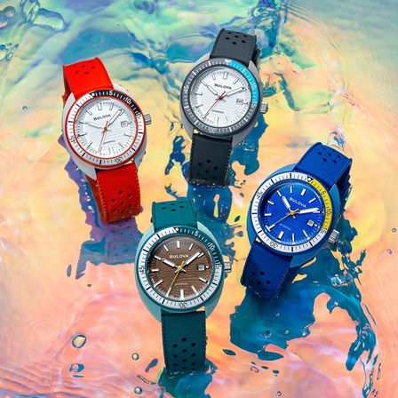 Vier Bulova-Uhren mit unterschiedlichen Armbandfarben, die in Wasser mit bunten Spritzern ausgestellt sind. Jede Uhr hat ein einzigartiges Zifferblattdesign.
