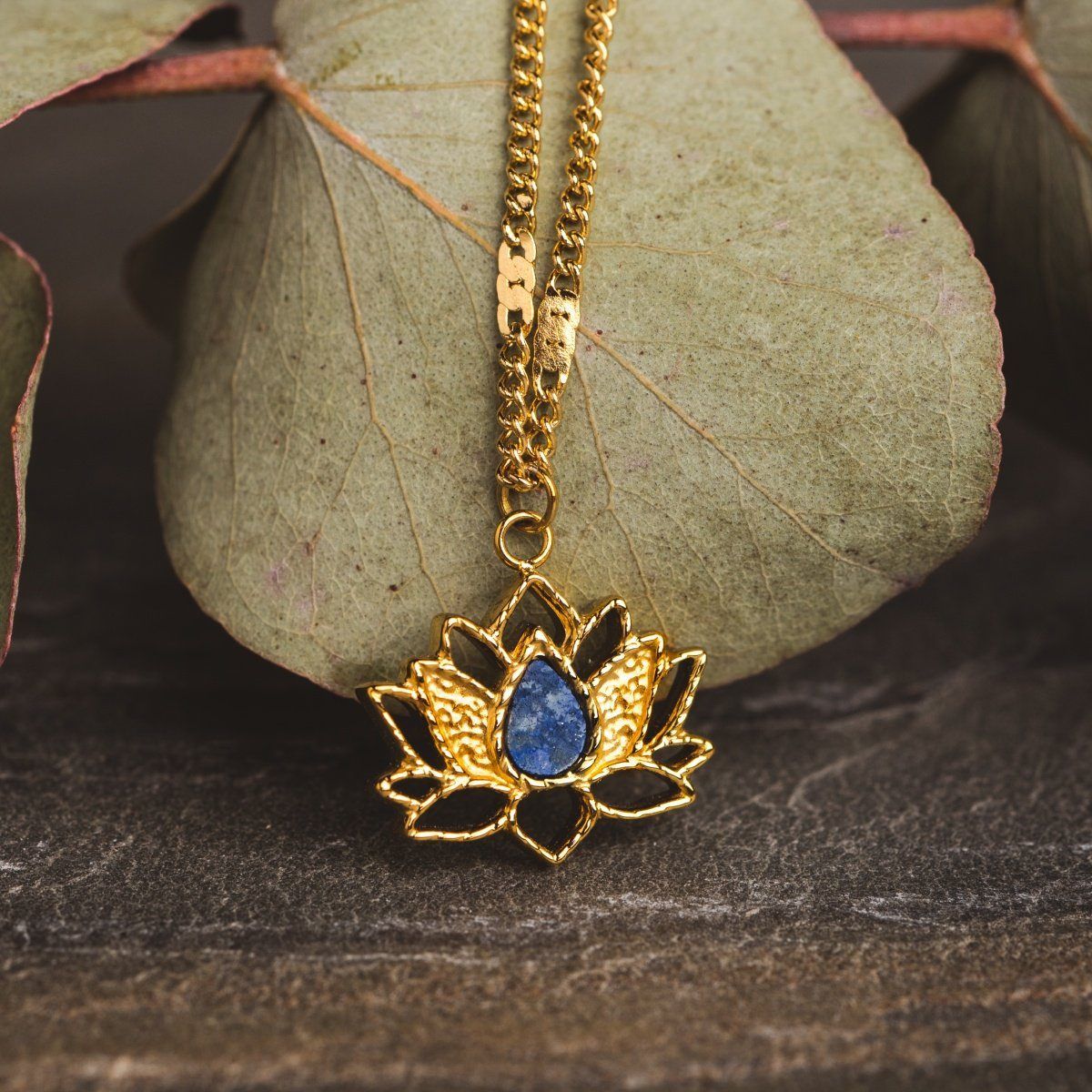 Eine goldene Halskette mit einem Lotus-Anhänger, der einen blauen Edelstein auf einem Blatthintergrund zeigt.
