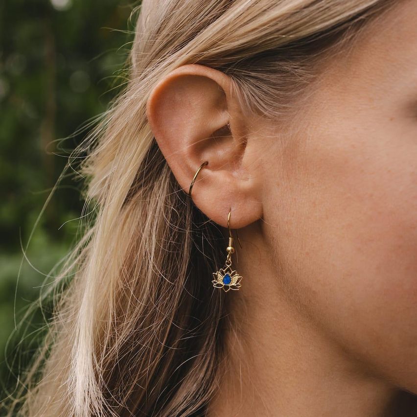Eine Frau mit langen blonden Haaren trägt goldene Lotos-Ohrringe mit blauen Steinen. Der Hintergrund ist verschwommen mit grünen Blättern.