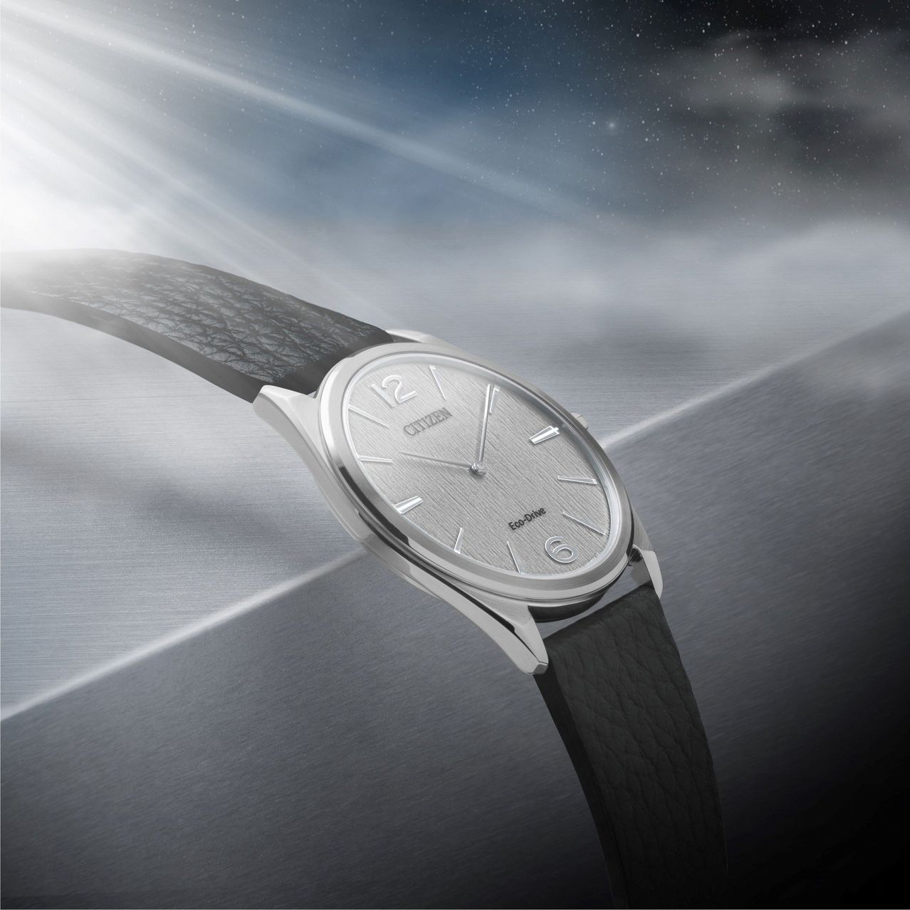 Eine silberne Armbanduhr mit einem schwarzen Lederarmband liegt auf einer metallischen Oberfläche mit einem bewölkten Nachthimmel im Hintergrund.