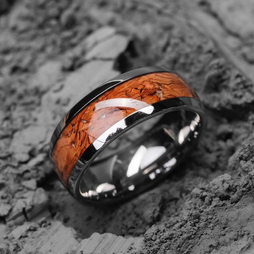 Ein schwarzer und silberner Ring mit einem Holzband liegt auf einer felsigen Oberfläche. Die Holzmaserung ist deutlich sichtbar.